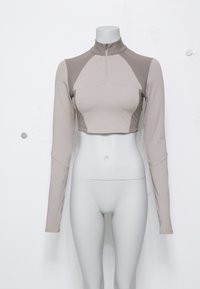 Lys beige cropped langermet topp med høy krage og halv glidelås. Har mesh-paneler på skuldrene og et glatt, elastisk stoff.