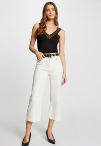 Canotta nera con bordo in pizzo, jeans bianchi a vita alta e cropped con orlo sfilacciato, e décolleté neri a punta. Cintura con fibbia oro come accento.
