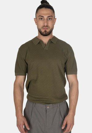 Uomo con capelli legati indossa una polo verde oliva a maniche corte texturizzata e pantaloni grigi, guarda avanti con espressione neutra.