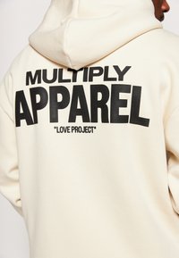 Sudadera de color crema con un gran estampado negro que dice "MULTIPLY APPAREL" y "LOVE PROJECT" en la parte trasera.