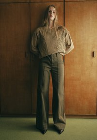 Beżowy sweter z koronkowymi detalami, zestawiony z oliwkowymi spodniami z szerokimi nogawkami i czarnymi czółenkami. W tle drewniana szafka na przechowywanie.