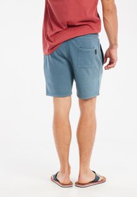Shorts bleus avec une coupe décontractée, une taille élastique et une poche arrière simple. Portés avec une chemise rouge et des tongs.