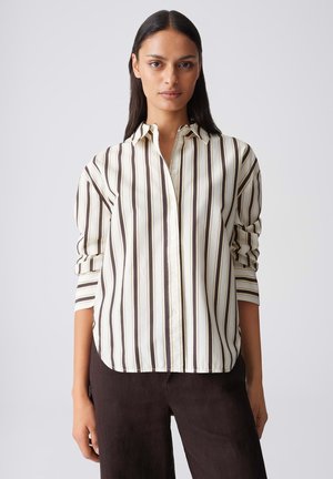 Donna che indossa una camicia bianca larga con bottoni, a righe verticali marroni e pantaloni scuri, in piedi davanti a uno sfondo chiaro e semplice.