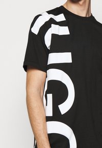 Mand iført sort T-shirt med kort ærme, hvor der er trykt store, fede hvide abstrakte bogstaver og former på fronten og ærmet.