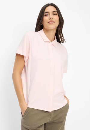 Frau mit langen dunklen Haaren, die ein hellrosa kurzärmeliges Hemd mit Kragen und olivgrüne Hosen trägt, die Hände in den Taschen, leicht lächelnd.