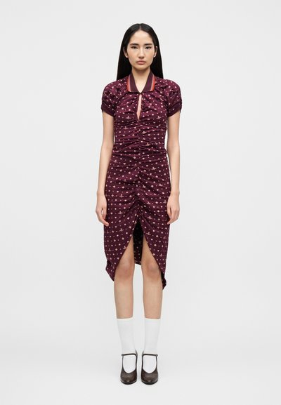 Femme debout portant une robe bordeaux froncée jusqu'aux genoux avec des manches courtes, décorée de petites fleurs beiges, associée à des chaussettes blanches et des talons marron.