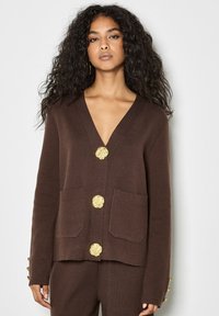 Cardigan in lana marrone con scollo a V, tre bottoni dorati a forma di fiore sul davanti e due tasche frontali. Tessuto morbido e testurizzato, vestibilità comoda.