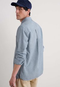 Világoskék, hosszú ujjú ing, amely chambray anyagból készült, felgombolt ujjakkal, hátsó redővel és ívelt alsó résszel, világos színű nadrággal párosítva.