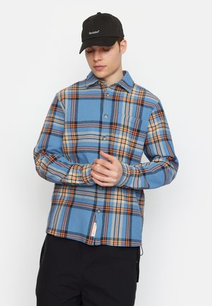 Blauw geruit flanellen shirt met oranje en gele accenten, lange mouwen, knoopsluiting aan de voorkant en een borstzak. Zwarte honkbalpet met logo.
