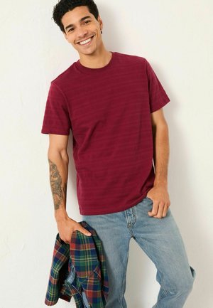 Gestreiftes T-Shirt in Kastanienbraun aus Baumwolle, kombiniert mit blauen Jeans. Das Modell hält ein grün-rotes kariertes Hemd. Einfaches Rundhaldesign.