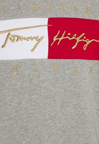 Szara bawełniana tkanina z poziomym wzorem i czerwoną oraz białą opaską. Na czerwonym pasku wyhaftowano złotymi literami napis "Tommy Hilfiger", poniżej znajduje się napis "M L X".