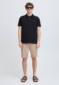 Camiseta polo negra de manga corta, pantalones cortos beige y sandalias marrones. La camiseta presenta un pequeño logo en el pecho.