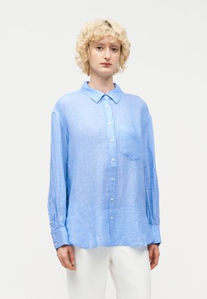 Osoba s krátkymi kučeravými blond vlasmi, ktorá nosí voľnú svetlomodrú košeľu na gombíky a biele nohavice, na jednoduchom pozadí.