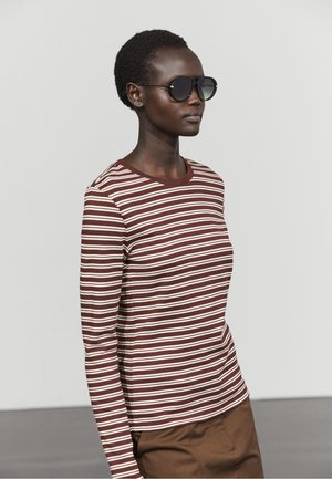 Woman wearing dark sunglasses, a brown and white striped long-sleeve shirt, and brown pants, facing right against a plain background.