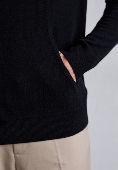 Svart strikkegenser med sidelomme, ribbestrikkede ermer og kant. Myk tekstur med en tettsittende silhuett. Et par lyse beige bukser synlige nedenfor.