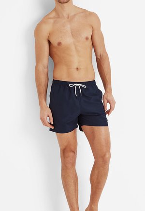 Shirtless man wearing dark navy swim trunks with a white drawstring stands against a plain white background.