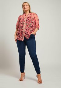 Blusa floreale in tonalità di rosa e viola, realizzata in un tessuto leggero, con chiusura a bottoni e maniche corte. Abbinata a jeans skinny blu scuro.