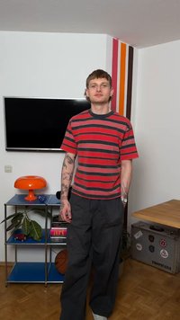 Νέος άνδρας με τατουάζ, φορώντας κόκκινη και μαύρη ριγέ μπλούζα και μαύρο παντελόνι, στέκεται σε ένα μοντέρνο δωμάτιο με τηλεόραση, πορτοκαλί λάμπα και ράφια με φυτά και βιβλία.