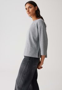 Femme avec de longs cheveux noirs portant un haut gris texturé à manches trois-quarts et un jean large taille haute gris foncé, le tout sur fond uni.