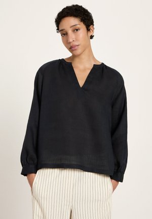 Woman with short curly hair wearing a loose black long-sleeve blouse and white pants with thin black vertical stripes.