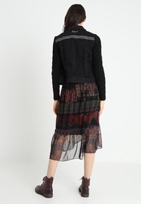 Nainen, jolla on päällään musta takki, jossa on teksturoidut hihat, kukkakuvioinen läpikuultava midi-hame ja tummanruskeat nilkkasaappaat, seisoo selkä kameraan päin.