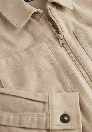 Bēšas krāsas rāvējslēdzēja jaka no mīksta auduma ar teksturētu virsmu, veidota ar apkakli, divām krūšu kabatām un tumšām pogu detaļām.