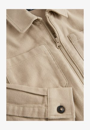 Jachetă bej cu fermoar, dintr-un material moale cu suprafață texturată, având guler cu rever, două buzunare la piept și nasturi închși în culoare închisă.