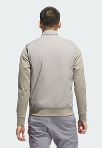 Lagani bež zip-up kaput s glatkom teksturom. Ima visoki ovratnik i elastičnu donju obrub. Nosi se preko majice s dugim rukavima i hlača.