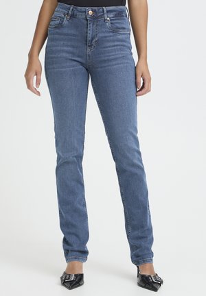 Kvinde iført tætsiddende mellemblå denimjeans og sorte spidse hæle, stående foran en ensartet lys baggrund.