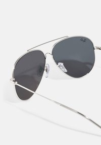 Ochelari de soare aviator cu lentile gri închis, ramă din metal argintiu și suporturi pentru nas transparente. Prezintă o bară subtire de sprijin și un design elegant.
