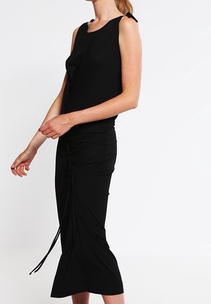 Femme portant un haut noir sans manches avec un nœud sur l’épaule et une jupe midi noire avec des détails à cordon.