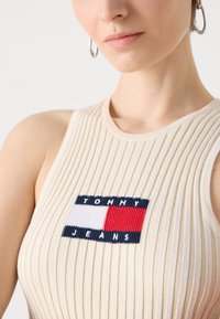 Bežinė raukšlėta maikutė su išsiuvinėtu "TOMMY JEANS" logotipu tamsiai mėlyna ir raudona spalvomis ant stačiakampio formos pleistro viduryje.