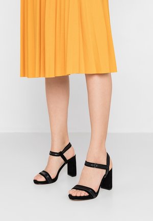 Jambes portant une jupe plissée jaune et des sandales noires à talons hauts avec des brides à la cheville sur un fond clair uni.