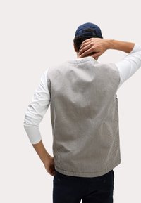 Gestreifte ärmellose Weste aus grauem und weißem Stoff, mit Rundhalsausschnitt und Rückentasche, getragen über einem langärmligen weißen Hemd.