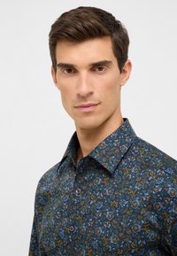 Uomo con capelli corti e scuri che indossa una camicia a bottoni con motivo floreale scuro su uno sfondo chiaro e uniforme.