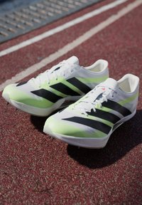 Chaussures de sport blanches et vert néon avec des rayures noires, dotées d'un design élégant et d'une tige texturée pour une meilleure adhérence, positionnées sur une surface de piste.