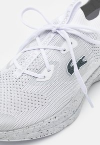 Sneaker en maille blanche avec une semelle texturée et tachetée, des lacets blancs et un logo crocodile noir sur le côté.