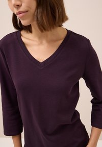 T-shirt viola con scollo a V e maniche a tre quarti. Il tessuto appare liscio con una leggera lucentezza. Dettaglio di cucitura sulla scollatura.