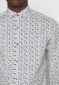 Biała koszula z guzikami i okrągłym granatowym wzorem. Posiada klasyczny kołnierzyk i gładką fakturę, z guzikami z przodu oraz detalami na mankietach.
