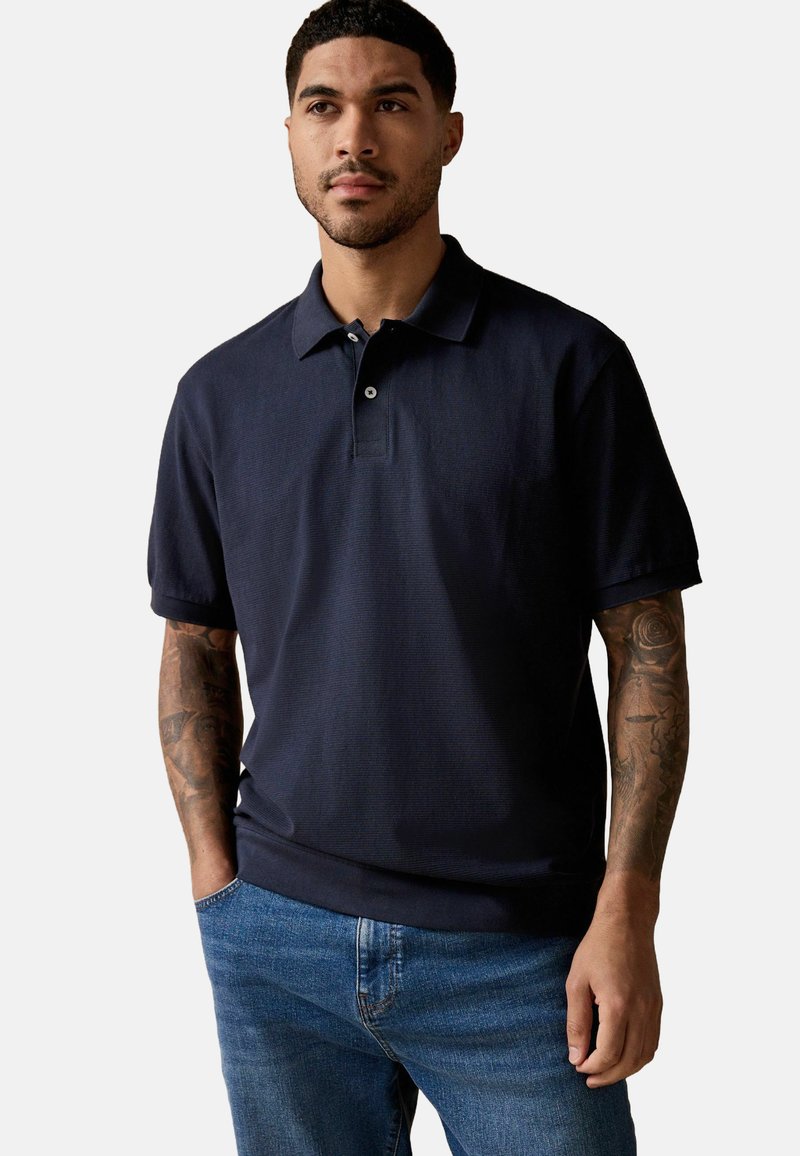 Marineblå polo skjorte lavet af struktureret stof med knaplukning, korte ærmer og klassisk krave, båret med blå jeans.