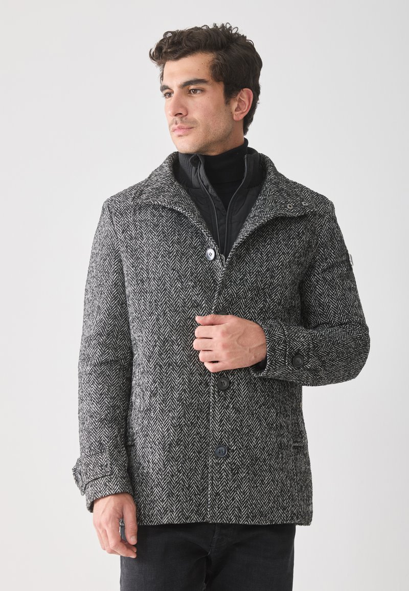 Hombre vestido con un abrigo de patrón espiga gris sobre una chaqueta negra con cremallera, de pie y mirando hacia un lado contra un fondo liso.