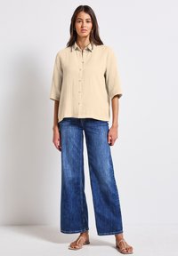 Camicia beige con bottoni, in tessuto testurizzato e maniche a tre quarti, abbinata a jeans in denim blu a gamba larga e sandali bassi.