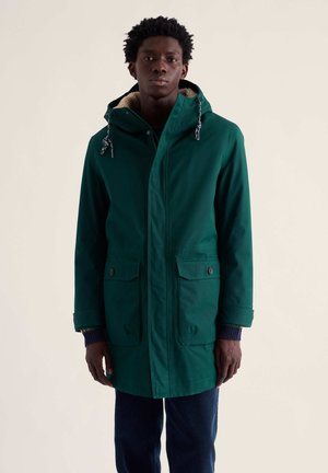 Young man wearing a green hooded winter coat with two front pockets, standing against a plain light background.