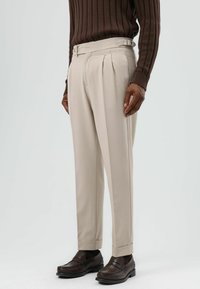 Beige plissé broek met zijwaartse verstelers, gemaakt van gladde stof. Gecombineerd met donkerbruine loafers. Getoond in staande positie.