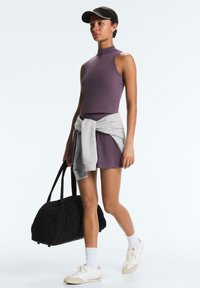 Femme en robe de sport violette sans manches et visière noire marche en portant un sac de sport noir, avec un sweat-shirt gris noué autour de la taille.