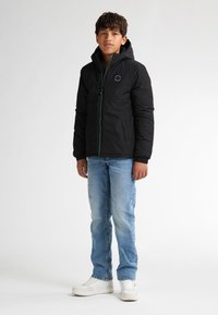 Giacca nera isolante con cappuccio, zip frontale, dettagli verdi e logo. Abbinata a jeans blu chiaro e scarpe da ginnastica bianche.