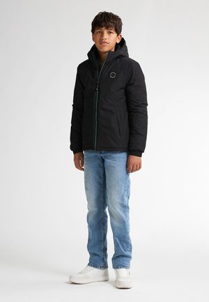 Schwarze isolierte Jacke mit Kapuze, Reißverschluss vorne, grünen Akzenten und Logo. Kombiniert mit hellblauen Jeans und weißen Sneakers.
