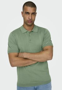 Polo shirt corto a maniche corte verde chiaro con colletto, patta con tre bottoni e polsini a coste, indossato con jeans in denim blu.