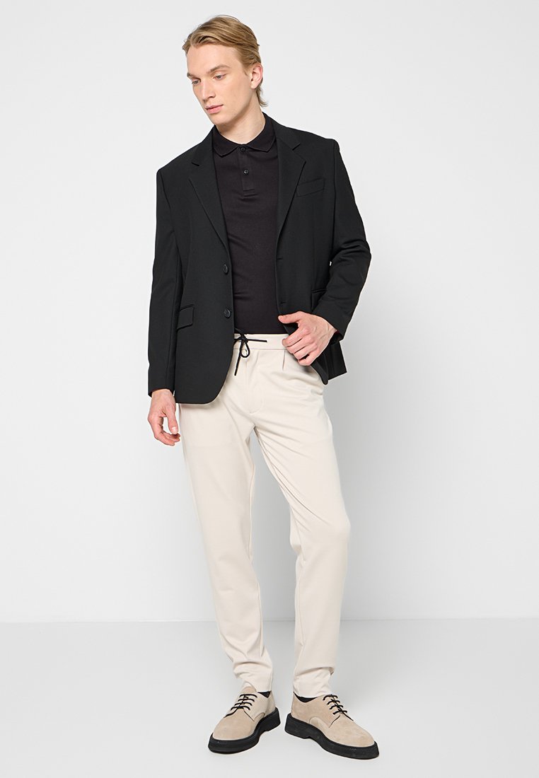Schwarzer Blazer über einem schwarzen Polo, kombiniert mit cremefarbenen Hosen und hellbraunen Schuhen. Glatte Materialien mit minimalistischem Design und ohne Muster.