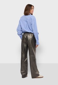 Lichtblauw gestreepte overhemdd met een rugplooitje, gecombineerd met glanzende metallic grijze wijde broek. Model staat met een gedraaid profiel.